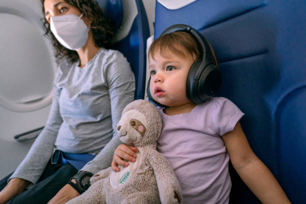 An adorable Eurasian toddler girl wears headphone and embraces a stuffed toy while watching a movie on an airplane. The child's mom is sitting next to her in the window seat and is wearing a protective face mask.